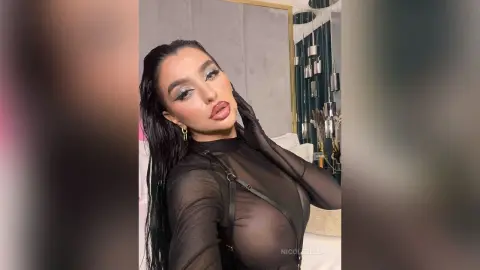 NicoleBella poses in a sheer black long-sleeve top with her dark hair wet-look and heavy eye makeup.