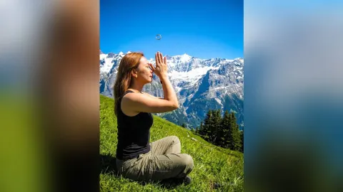 MilaSwan — MilaSwan sits cross-legged on a grassy mountain slope with her eyes closed and hands pressed together in prayer.