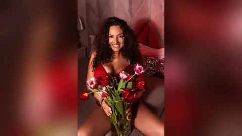 AishaApril smiles while holding a large bouquet of red and white tulips against her chest, wearing a red lace bodysuit or lingerie set.