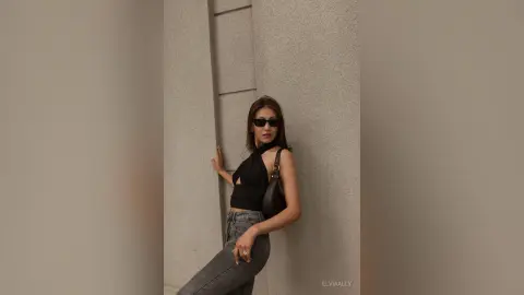 Stylish woman in a black halter top and grey jeans posing against a minimalist concrete wall.