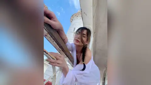 Young woman in a white blouse leaning on a balcony railing against a bright blue sky background.