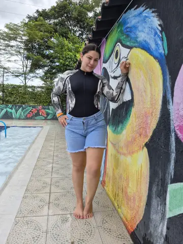 Smiling woman in a black patterned zip-up top and denim shorts posing by a colorful parrot mural near a pool.
