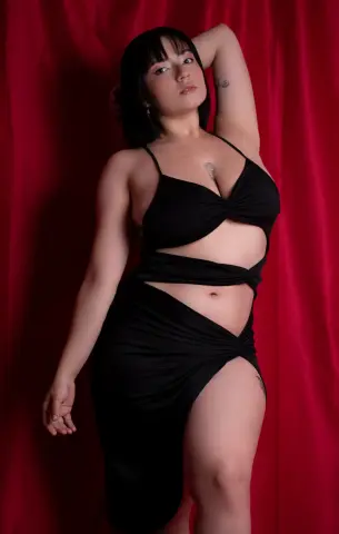 Woman in a black cutout maxi dress posing against a red velvet curtain in a moody studio setting.