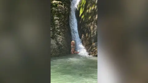 Blonde woman in a bikini relaxing by a tropical waterfall in a lush jungle lagoon.