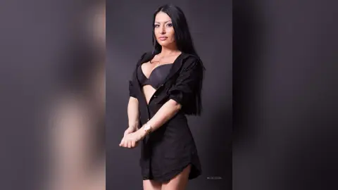 NicolleSun poses in a black bra and an open black button-down shirt, showing her long black hair and slim figure against a dark background.