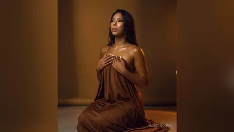 Elegant woman in a flowing brown satin dress posing gracefully in a warm, moody studio setting.