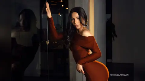 MarcelaMilan poses in a tight, off-the-shoulder brown dress with her long dark hair flowing down.