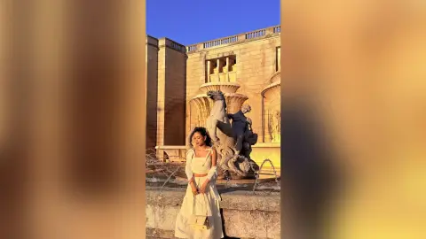 Young woman in a white summer outfit standing before a classical stone fountain in warm golden hour sunlight.