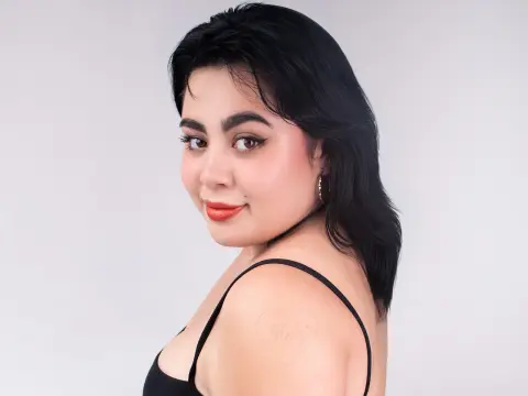 Smiling young woman with dark hair and red lipstick wearing a black tank top in a bright studio setting.