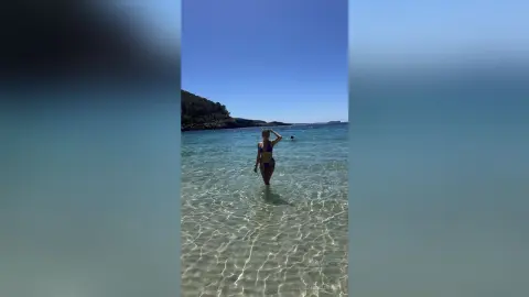 Young woman in a dark bikini standing in crystal clear turquoise ocean water under a bright sunny sky.