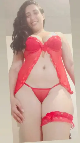 Smiling woman in red lace lingerie and matching garter set posing in a bright indoor setting.