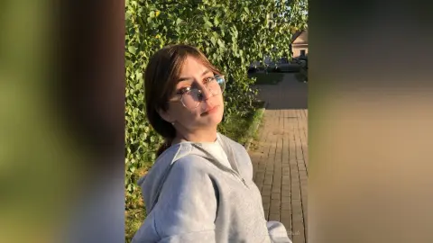 Young woman in a grey sweatshirt smiling outdoors near lush green trees in soft sunlight.