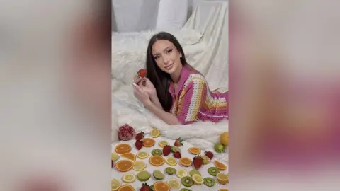 Smiling woman in a pink crochet cardigan enjoying fresh strawberries on a white fuzzy bed setting.