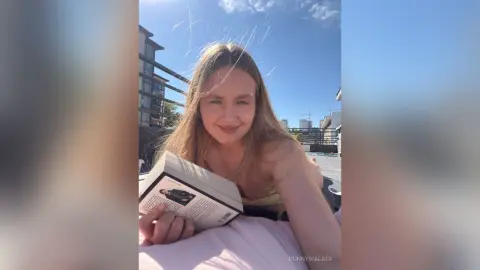 BonnyWalace smiles while lying on a pink pillow outdoors under a blue sky, holding a book in her hands.