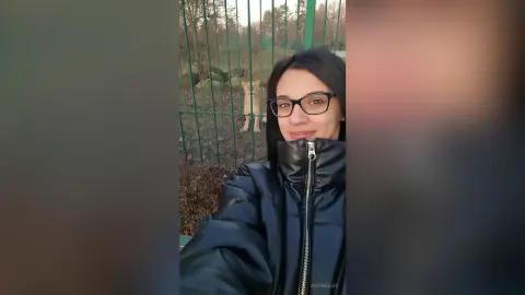 Smiling young woman in a black puffer jacket standing outdoors near a metal fence on a cloudy day.