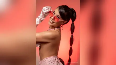 SamanthaHarvey smiles while wearing pink sunglasses, light pink gloves, and a pink lace outfit against a red background.
