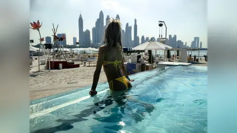 Blonde woman in a yellow bikini relaxing in a rooftop infinity pool overlooking the Dubai skyline at sunset.