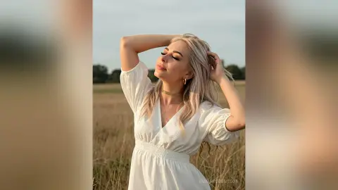Blonde woman in a white summer dress posing peacefully in a golden field at sunset.