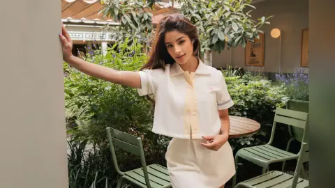 TefyRusso poses outdoors in a white short-sleeved shirt and matching skirt, leaning one arm against a wall surrounded by green plants and garden furniture.