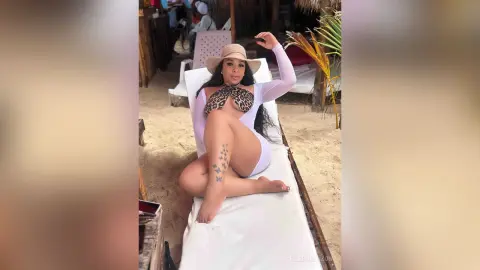 Smiling woman in a white cutout swimsuit and sun hat relaxing on a lounge chair at a tropical beach resort.
