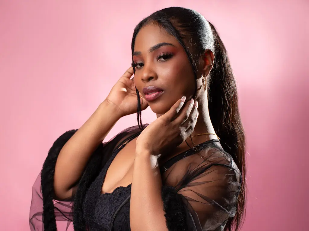 ZendayaRosse poses with her hands against her face, wearing a black lace top and sheer black sleeves against a pink background.