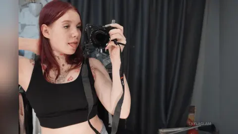 Redheaded woman in a black crop top taking a playful mirror selfie with a professional DSLR camera.