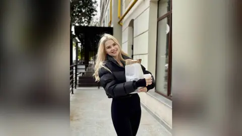 Smiling blonde woman in a black puffer jacket and leggings holding coffee and a bag on a city street.