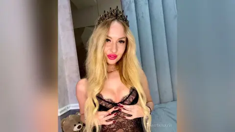 Blonde woman in black lace lingerie and a dark crown posing glamorously in a soft-lit bedroom setting.