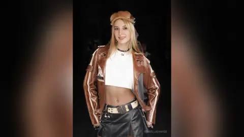 Blonde woman in a brown leather jacket and black skirt posing confidently in a dark setting.