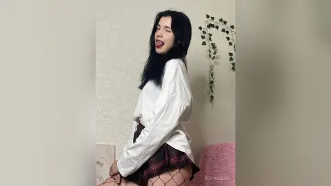 Playful young woman in a white oversized shirt and plaid skirt posing in a minimalist room with ivy decor.
