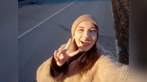 KailaDuch — Smiling young woman in a cozy beige teddy coat and knit beanie making a peace sign on a sunny road.