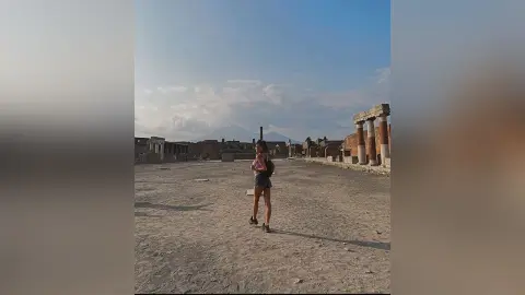 AnaMathers stands in an open outdoor ruins setting wearing a dark short romper or bodysuit.