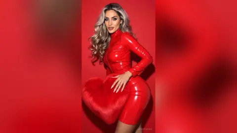 JessycaPage poses in a tight red latex outfit while holding a large red furry heart against a red background.