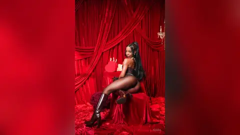 Glamorous woman in black bodysuit and boots sitting against red velvet curtains in a moody, romantic setting.