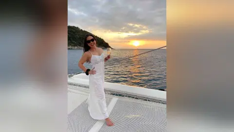 Smiling woman in a white sundress holding a drink on a boat at sunset over the ocean.