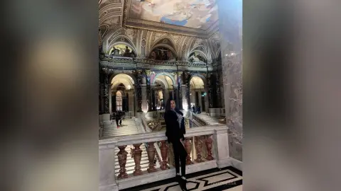 Stylish woman in a black outfit standing in a grand, ornate marble palace with classical architecture.