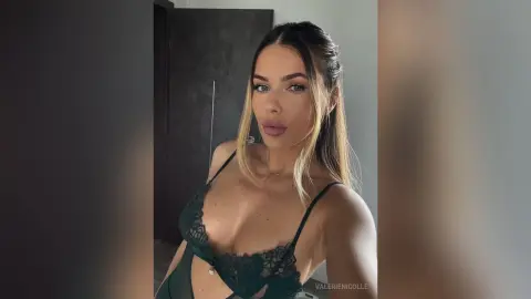 Young woman in a dark green lace bodysuit posing for a glamorous selfie in a modern indoor setting.
