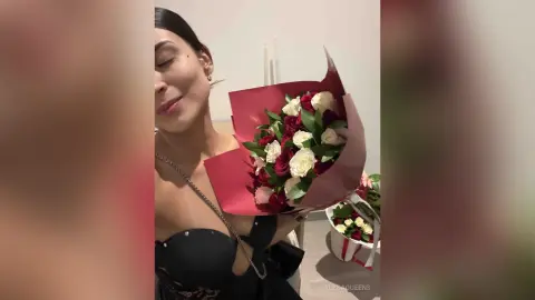 Smiling woman in a black dress holding a large bouquet of red and white roses in a cozy indoor setting.