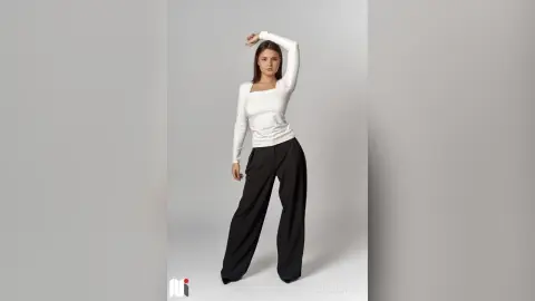 CaritaMattox poses in a white long-sleeve shirt and black wide-leg pants against a plain background.
