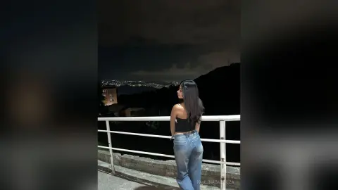 Young woman in a black tank top and blue jeans standing on a balcony overlooking a city at night.