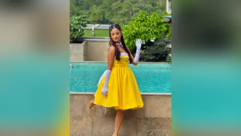 TefyRusso poses in a bright yellow dress and long white gloves while standing next to a swimming pool surrounded by green trees.
