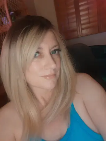 Smiling blonde woman in a blue tank top relaxing in a cozy indoor setting.