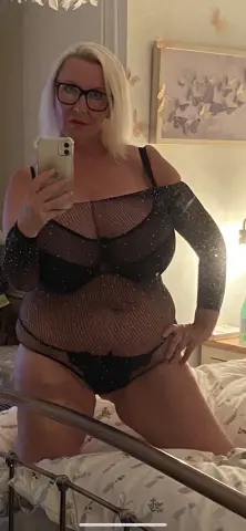 Blonde woman in black sheer mesh lingerie posing in a bedroom setting with a confident mood.