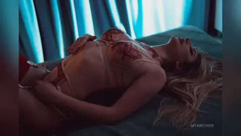 Blonde woman in sheer lace lingerie relaxing on a bed in a moody, blue-toned bedroom setting.