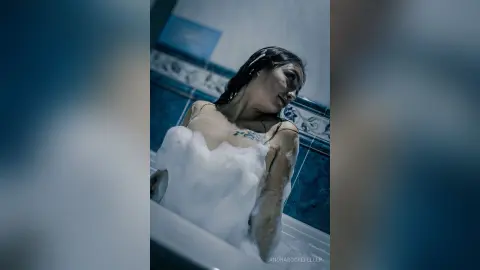 AnoraRockefeller with wet dark hair relaxing in a bubble bath, showing her bare chest and shoulders against blue tiled walls.