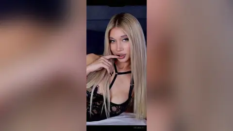 Blonde woman in a black lace halter top posing with a soft, intimate expression indoors.