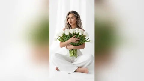 Elegant woman in white linen outfit holding a large bouquet of white tulips in a bright, minimalist setting.