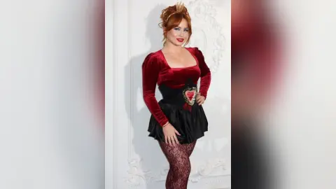 Redheaded woman in a vintage velvet burgundy and black dress posing elegantly in a bright white room.