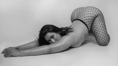 KimRoberth arches her back while lying on her stomach on a plain white floor, wearing black fishnet tights and black bottoms.