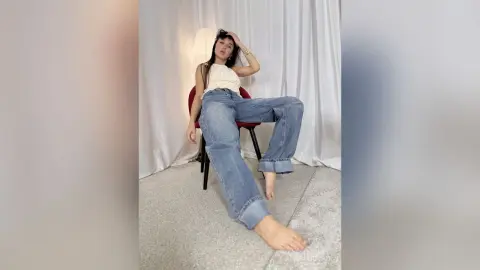 Young woman in a white tank top and baggy blue jeans sitting relaxed on a chair in a bright studio setting.